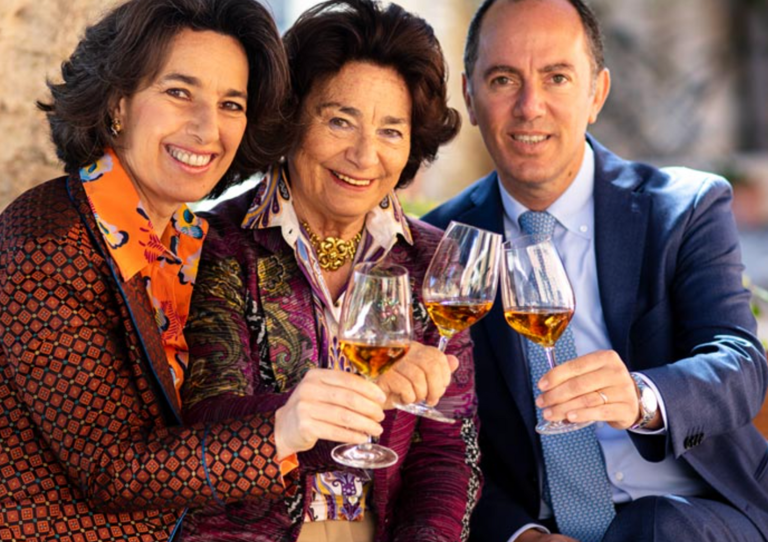 Drei elegant gekleidete Personen stoßen draußen fröhlich mit Weißweingläsern an.