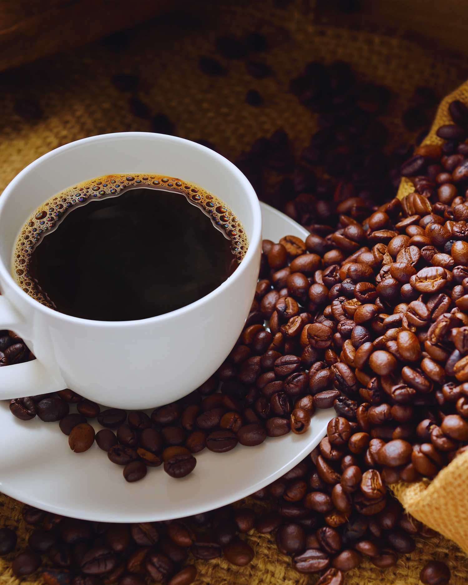 „Tasse schwarzer Kaffee auf Untertasse, umgeben von gerösteten Kaffeebohnen auf Jutesack“