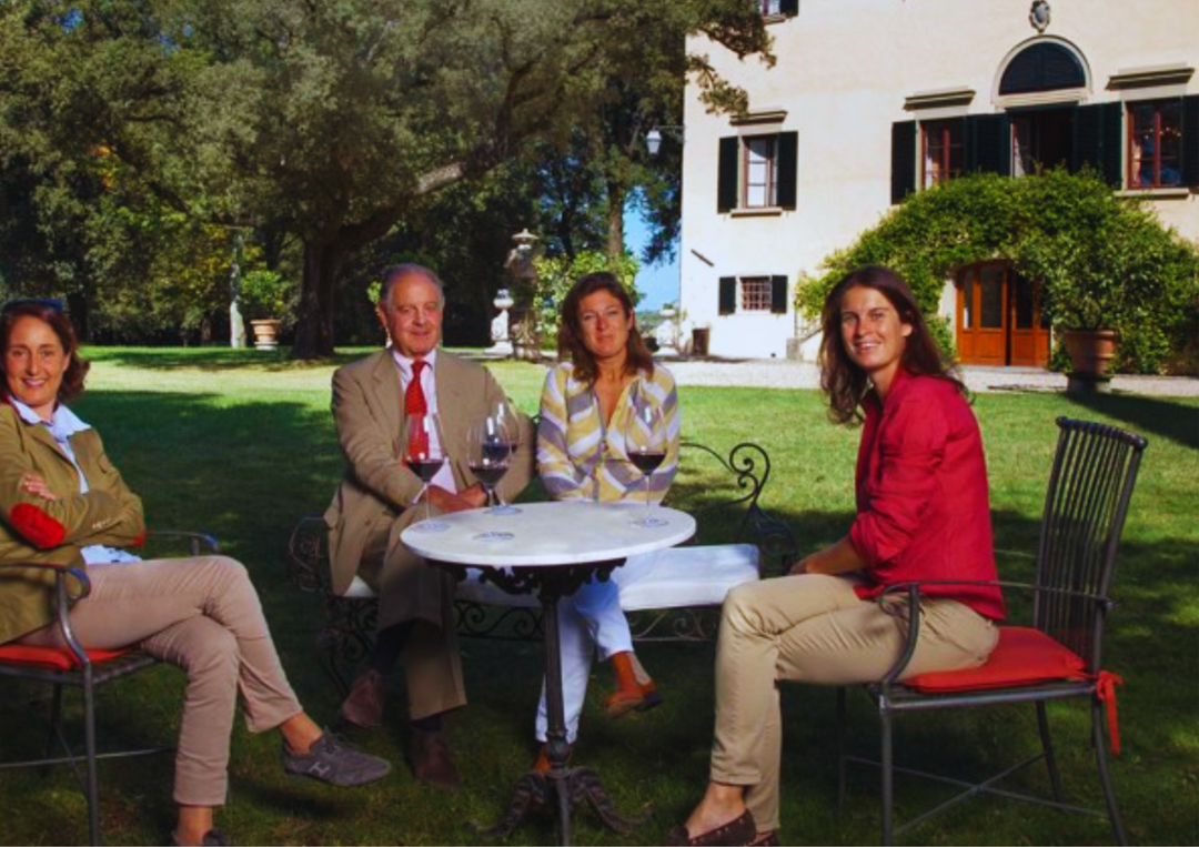 Vier Personen sitzen im Garten an einem runden Tisch vor einem großen Haus und genießen Wein.