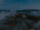 Kleines Dorf auf einem Hügel, umgeben von Weinbergen, teils von morgendlichem Nebel umhüllt.