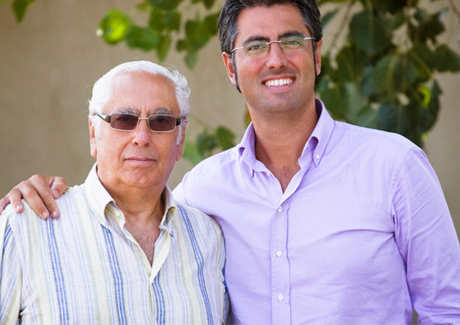 Zwei Männer, Arm in Arm, lächeln gemeinsam im Freien vor grünen Blättern.